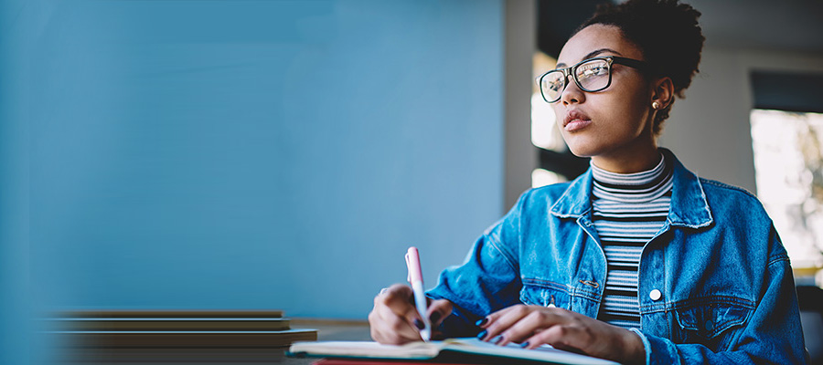 A young woman with curly hair and glasses is writing in a notebook wearing a denim jacket and striped turtleneck.