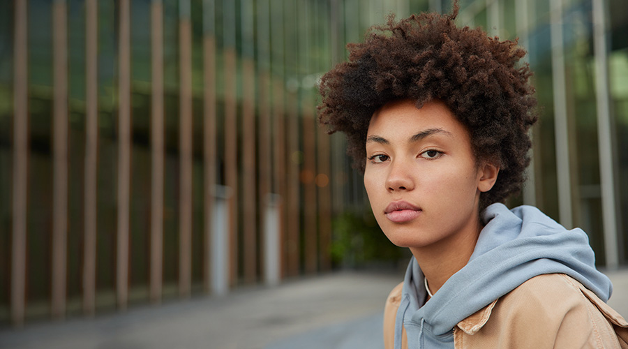 A young person with curly hair, wearing a gray hoodie and tan jacket, gazes into the camera