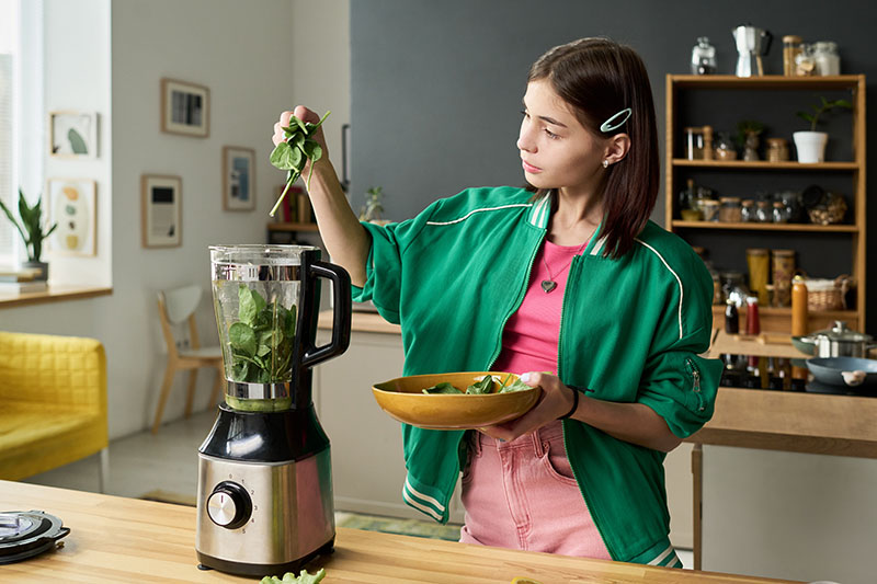A girl in a green jacket adding spinach to a blender in a bright kitchen.