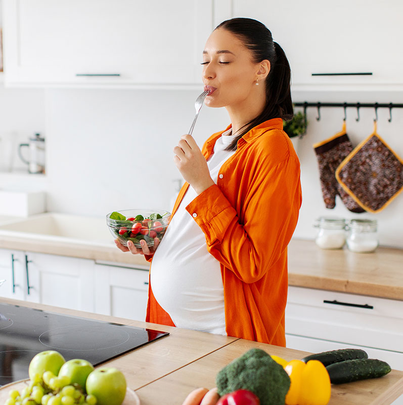 Pregnant woman in an orange dress enjoying a salad in a modern kitchen.