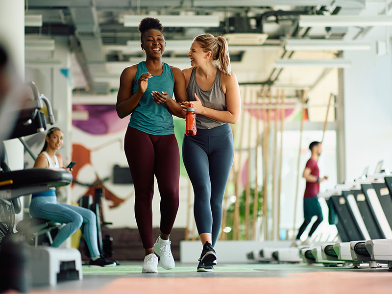 Two women walking and smiling in a gym, with exercise equipment and other people in the background.