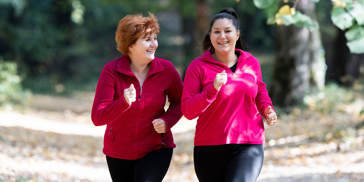 Two women are jogging together in a park, both smiling and wearing matching red jackets.