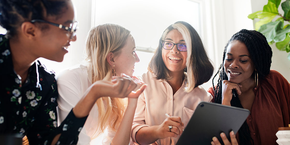 A diverse group of four women is gathered around a table, smiling and engaged in a discussion while one woman holds a tablet.