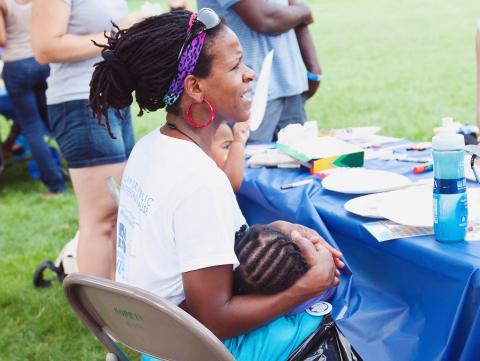 A caregiver sits on a chair outdoors, holding a child with braided hair, surrounded by art supplies on a table and fellow participants.