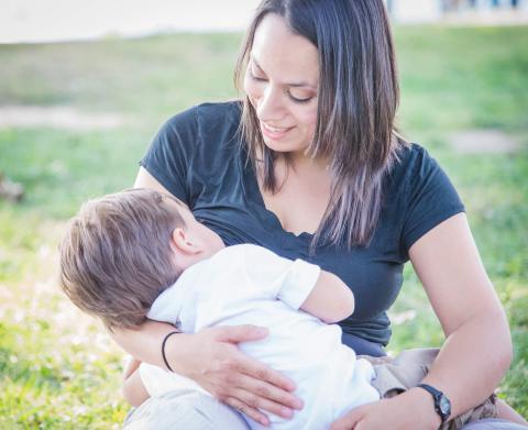 A woman in a black shirt cradles a young child in her arms while sitting on grass in a sunlit outdoor setting.