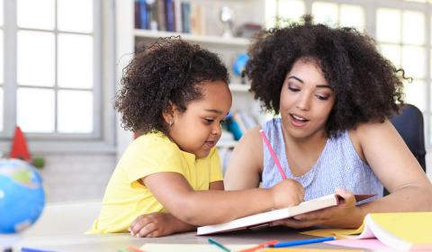 A woman and a girl with curly hair sit at a table, engaging in a creative activity with a book and colored pencils. Bookshelves and windows in the background.
