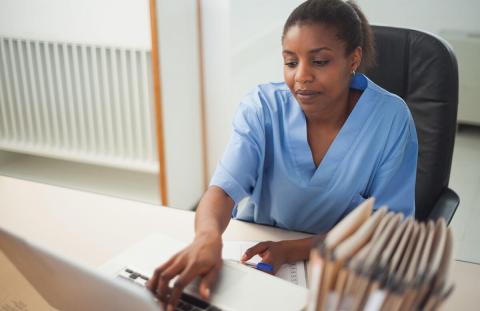 A healthcare professional in a blue uniform sits at a desk using a laptop, with folders stacked on the side.