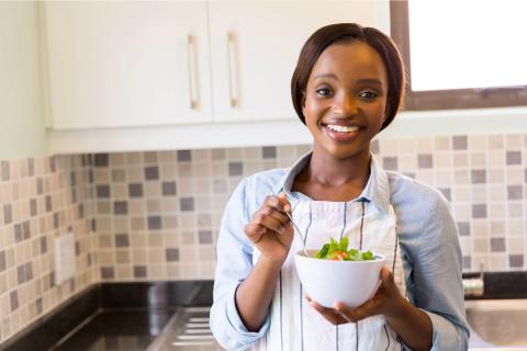 A person in a kitchen holds a bowl of fresh salad, preparing to enjoy a healthy meal. The kitchen features a stylish tile backsplash.