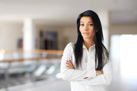 A woman with long black hair stands confidently with arms crossed, wearing a white shirt, in a bright, modern indoor space.