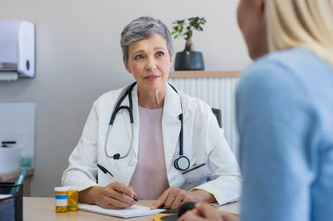 A doctor in a white lab coat sits at a desk with a stethoscope, discussing something with a patient. Prescription bottles are visible.