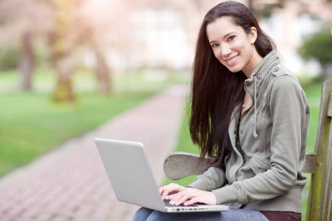 A young woman sits on a bench outdoors, focused on her laptop in a lush green park, with a pathway blurred in the background.