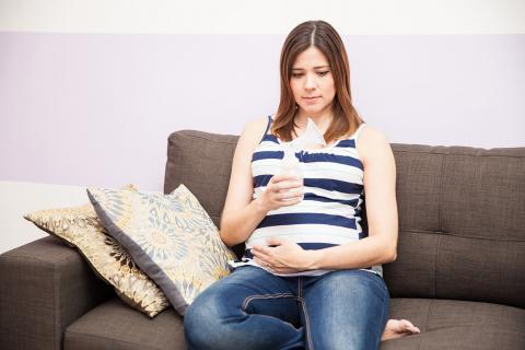 A pregnant woman sits on a couch, holding a bottle and cradling her belly, surrounded by patterned pillows and soft decor.