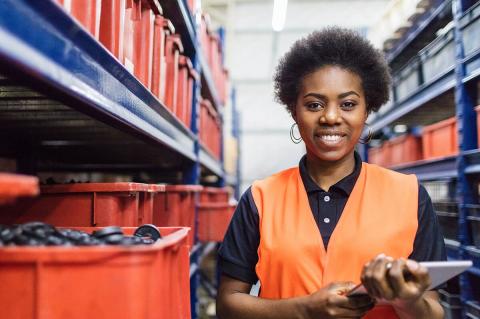 A worker in an orange vest stands in a warehouse aisle with red storage bins, holding a tablet and surrounded by shelves.