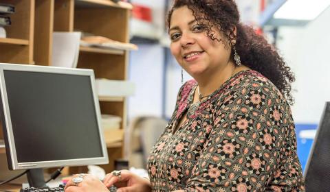 A person wearing a patterned dress sits at a desk with a computer, focused on their work in a cozy office environment.