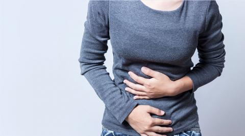 A person in a gray long-sleeve shirt clutches their stomach, indicating discomfort or pain against a plain light background.