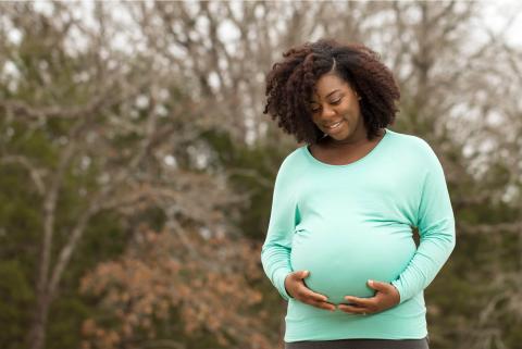 A pregnant woman stands outdoors, gently cradling her baby bump, wearing a light blue top against a blurred natural backdrop.