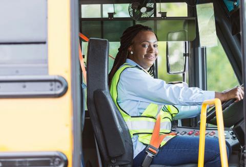 A woman wearing a blue shirt and high-visibility vest operates a bus, seated behind the wheel with safety features visible.