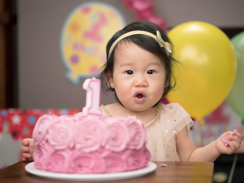 A delighted child in a decorative dress holds a piece of pink frosting cake at a birthday celebration, with balloons in the background.