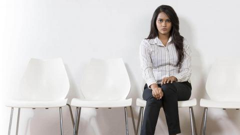 A woman sits on a chair, dressed in a striped shirt and black pants, with empty white chairs beside her against a plain wall.
